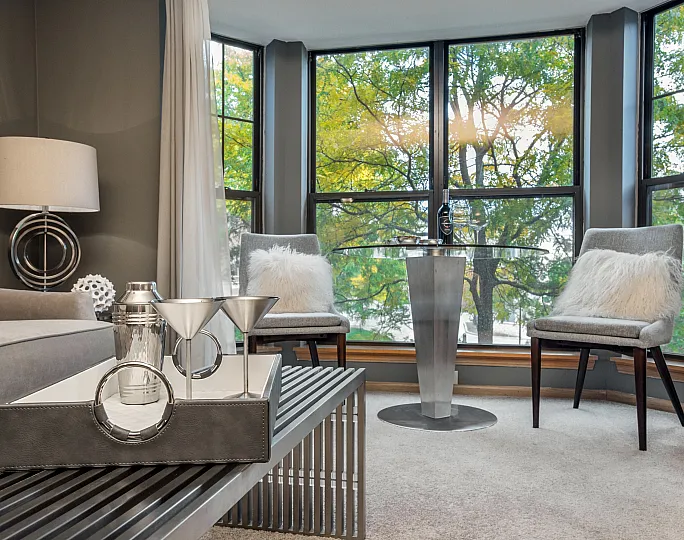 Elegant living room with modern decor, featuring large windows with lush green views, cozy seating, and a sophisticated metal table.
