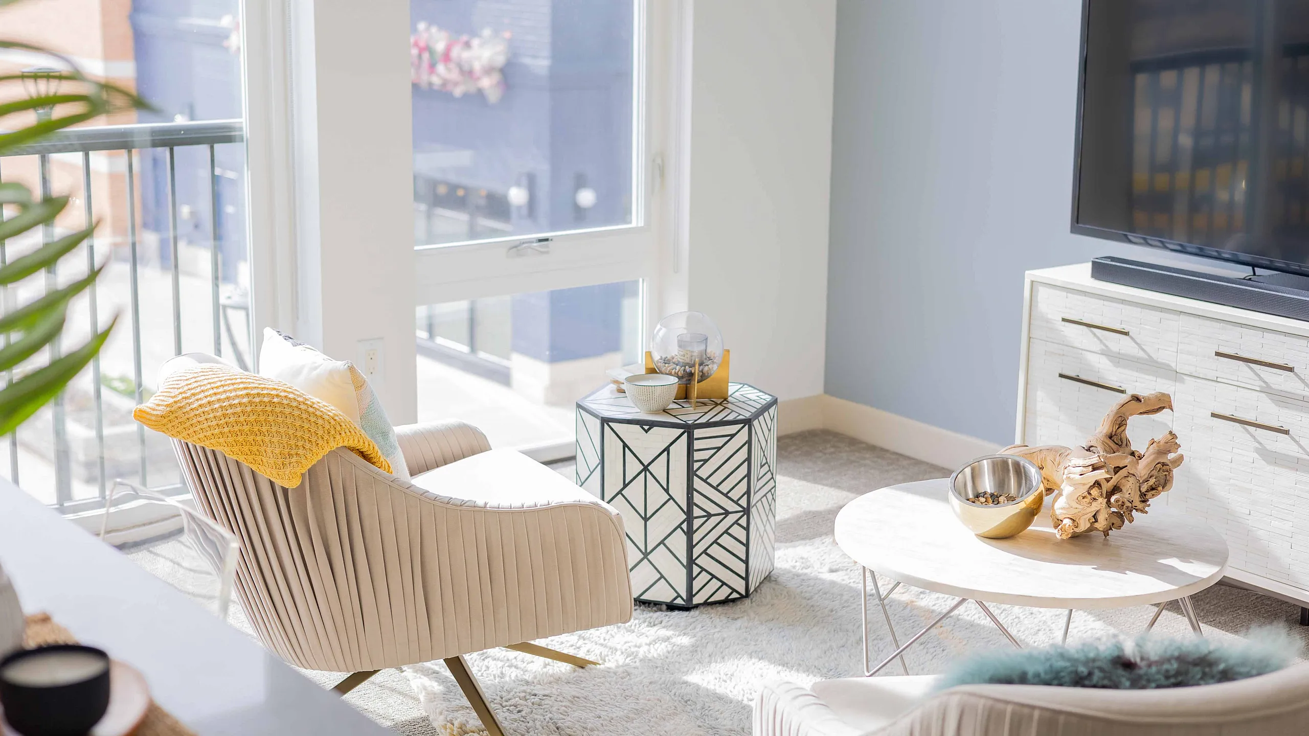 Bright and inviting living room with modern chairs, stylish side table, and large flat-screen TV; ample natural light streaming through expansive windows.