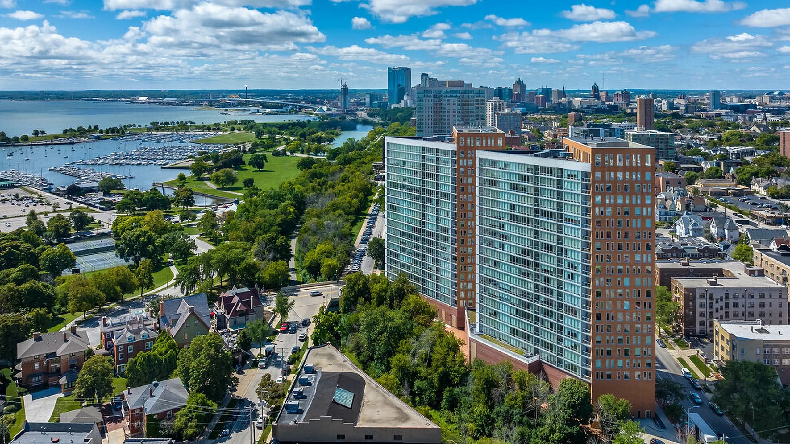 Breathtaking aerial view of a luxurious high-rise apartment complex overlooking a picturesque marina and vibrant city skyline, set against a backdrop of lush greenery and blue skies.