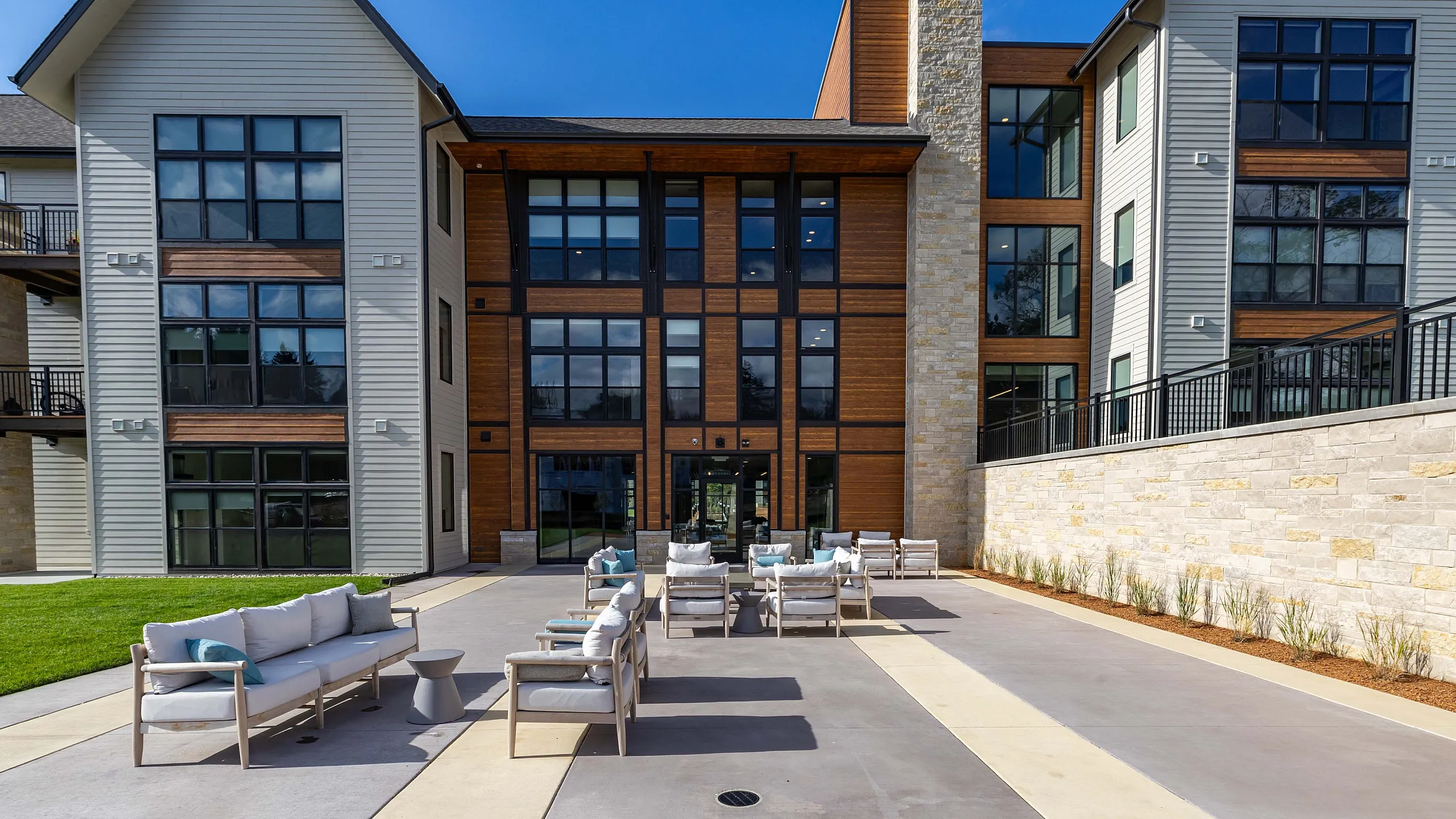 Modern apartment building featuring expansive glass windows and stylish outdoor patio with comfortable seating, ideal for relaxation and social gatherings, under a clear blue sky.