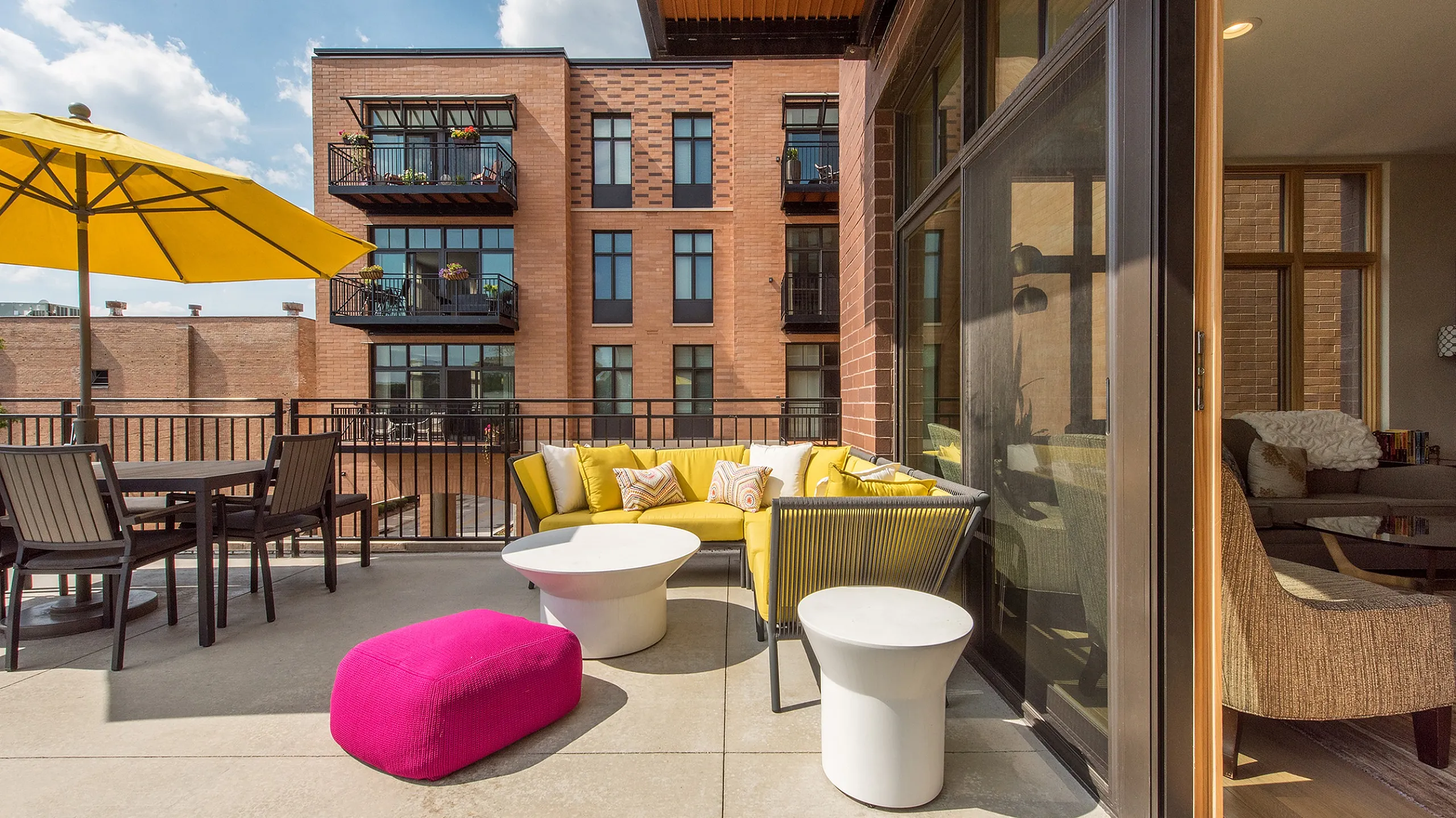 Elegant urban apartment with a stylish balcony featuring vibrant yellow seating, a pink ottoman, and an inviting outdoor dining area under a sunny yellow umbrella.