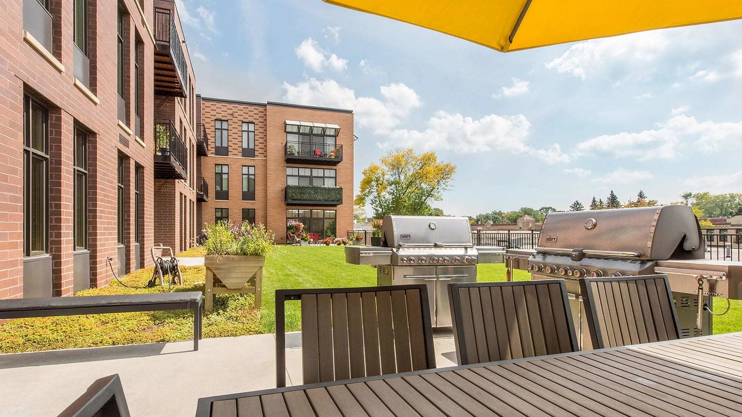 Modern apartment patio featuring sleek dining area, dual grills, and lush green space under a sunny yellow umbrella.