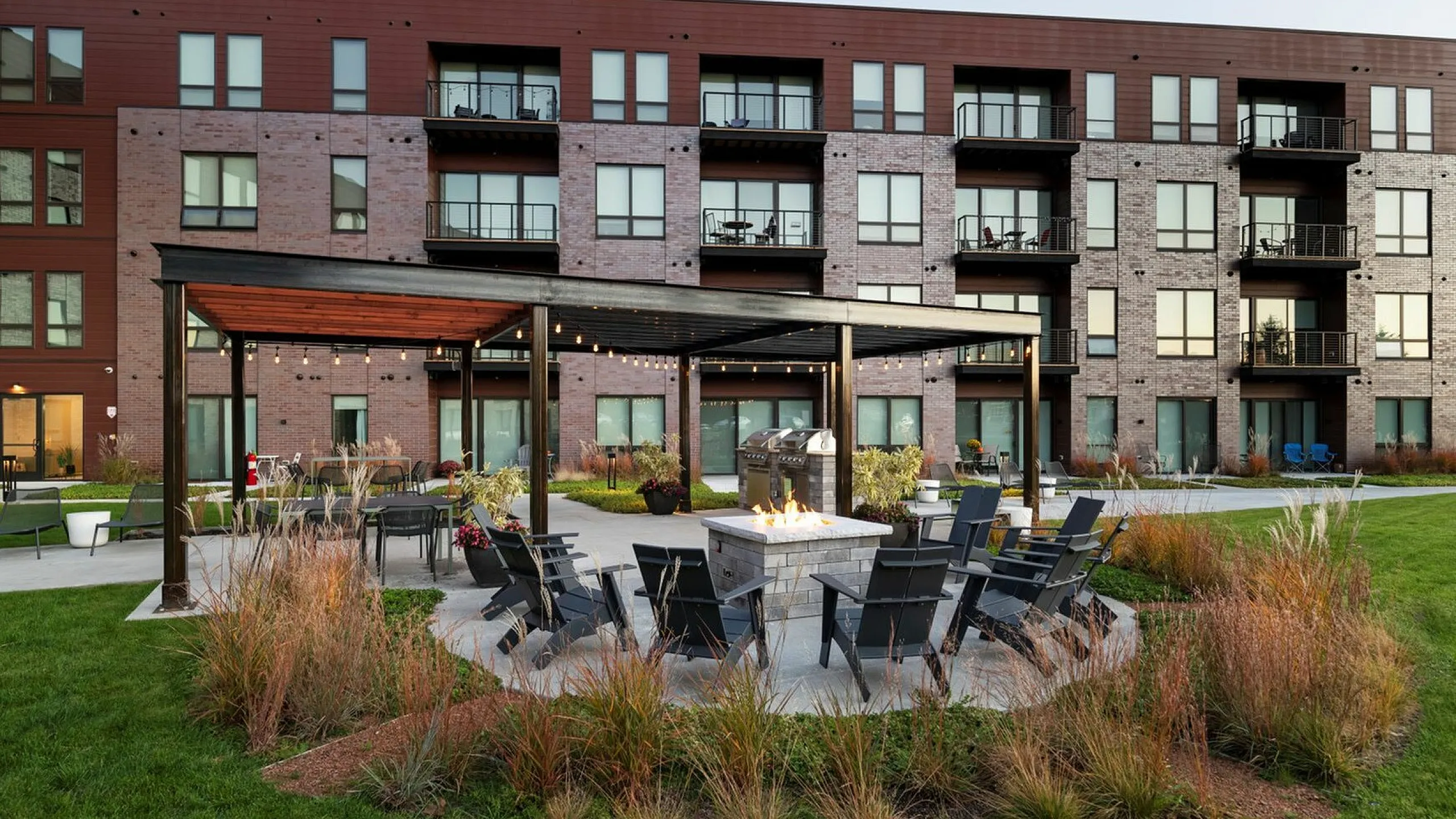 Charming apartment complex with modern brick facade features a lush green courtyard, stylish pergola, and inviting fire pit area for outdoor relaxation.