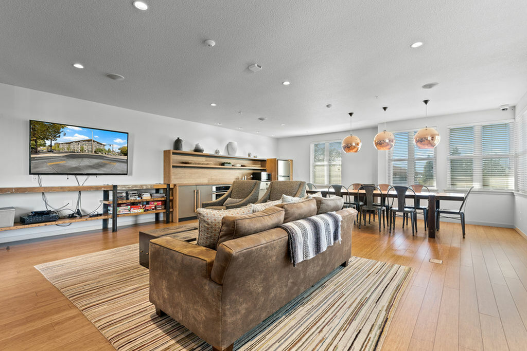 Bright and spacious community room with modern decor featuring a brown leather sofa, sleek wooden flooring, and large dining area with pendant lights.