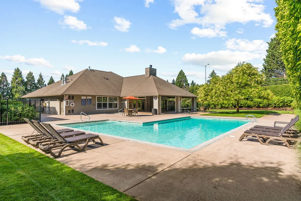 Charming outdoor pool area with inviting sun loungers and a cozy clubhouse surrounded by lush greenery and a clear blue sky.