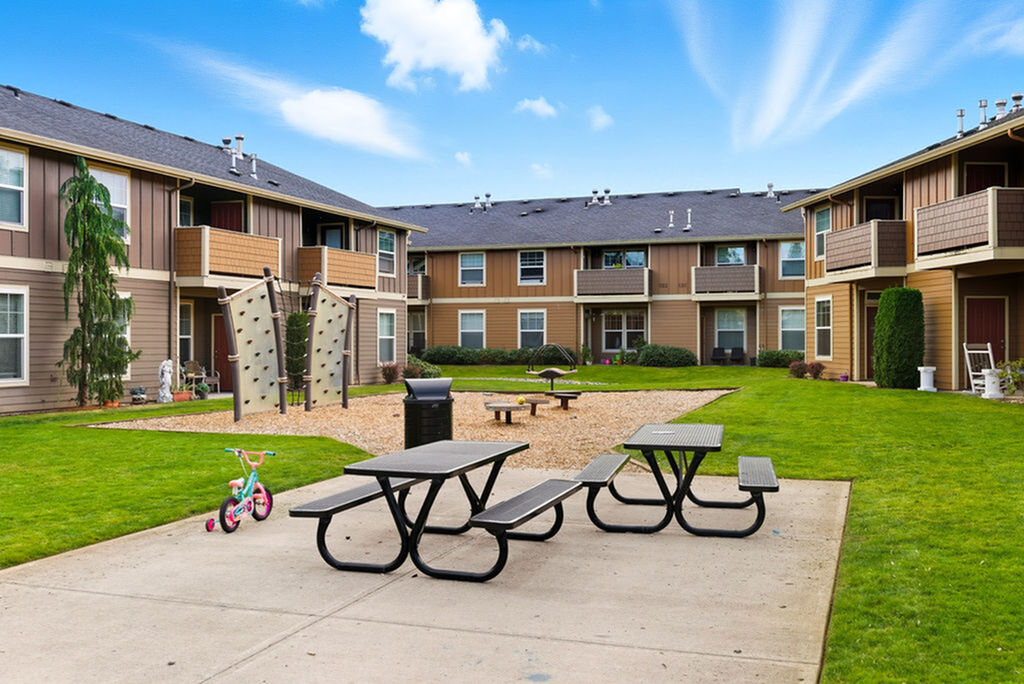Bright, family-friendly apartment complex courtyard featuring picnic tables, children\'s playground, and lush landscaping.
