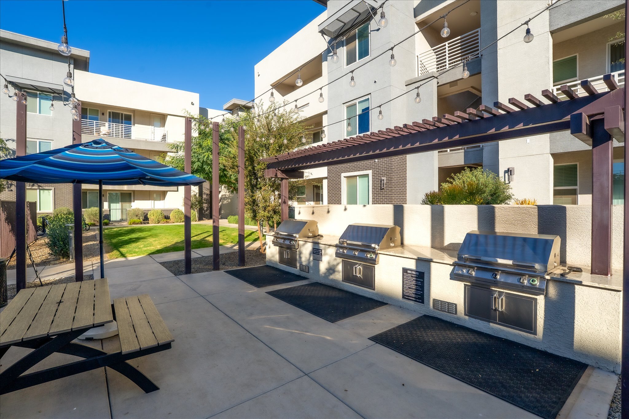 Chic outdoor communal area with modern BBQ grills, shaded picnic table, and lush landscaping, perfect for entertaining and relaxation.