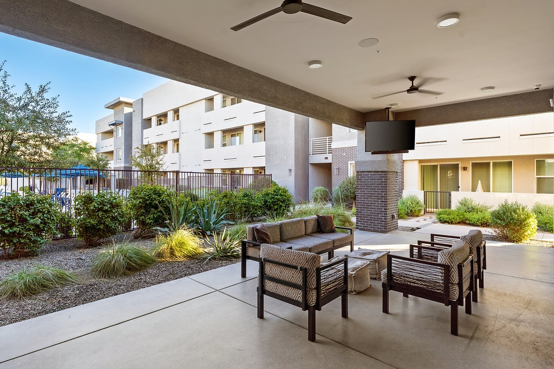 Modern open-air patio with cozy seating, ceiling fans, and a mounted TV overlooking lush greenery and a stylish apartment exterior.