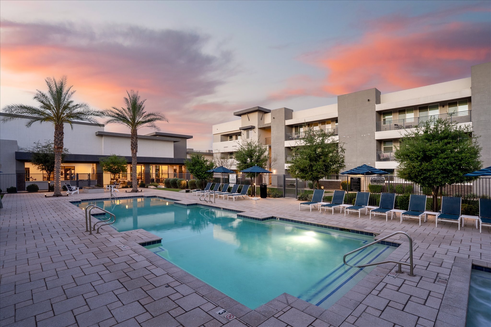 Elegant apartment complex with a luxurious swimming pool and sun loungers surrounded by lush greenery and modern architecture under a stunning sunset sky.