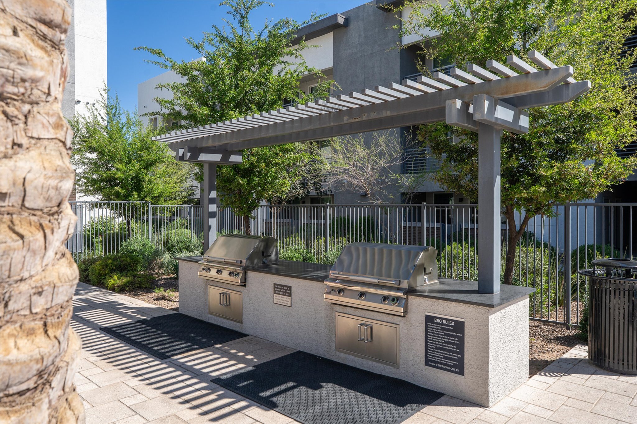 Modern outdoor grilling station with stainless steel grills and a sleek pergola, surrounded by lush greenery in a stylish apartment complex.