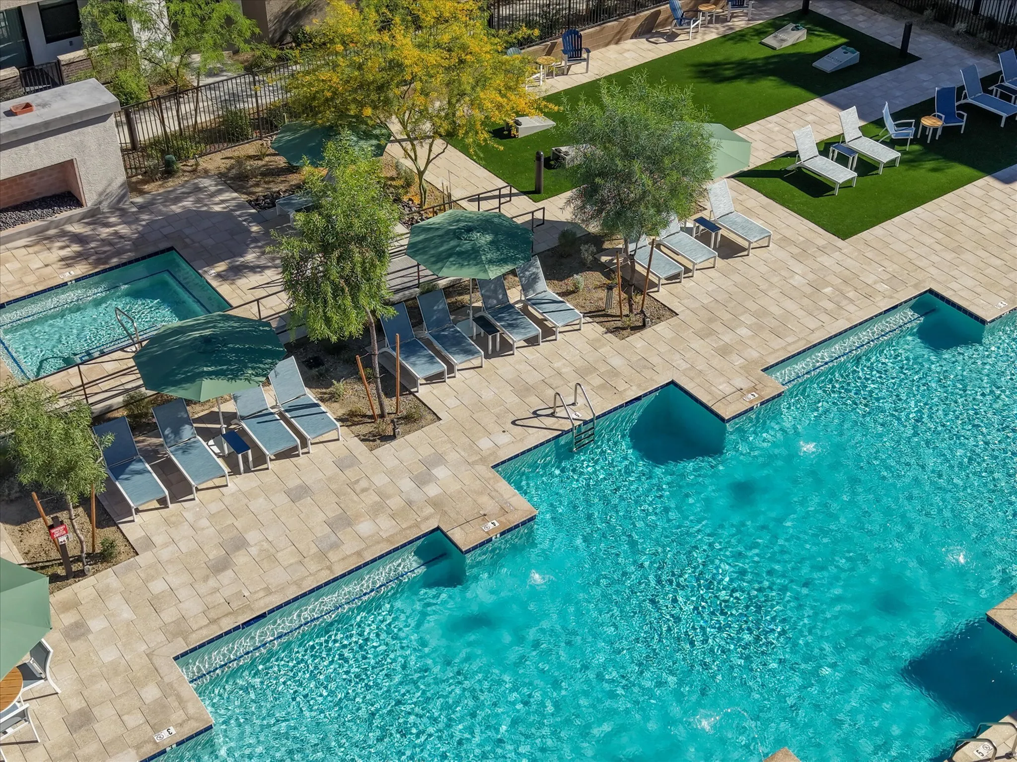 Luxurious apartment pool area featuring sparkling swimming pool, cozy hot tub, and stylish lounge chairs under verdant umbrellas in a lush green setting.