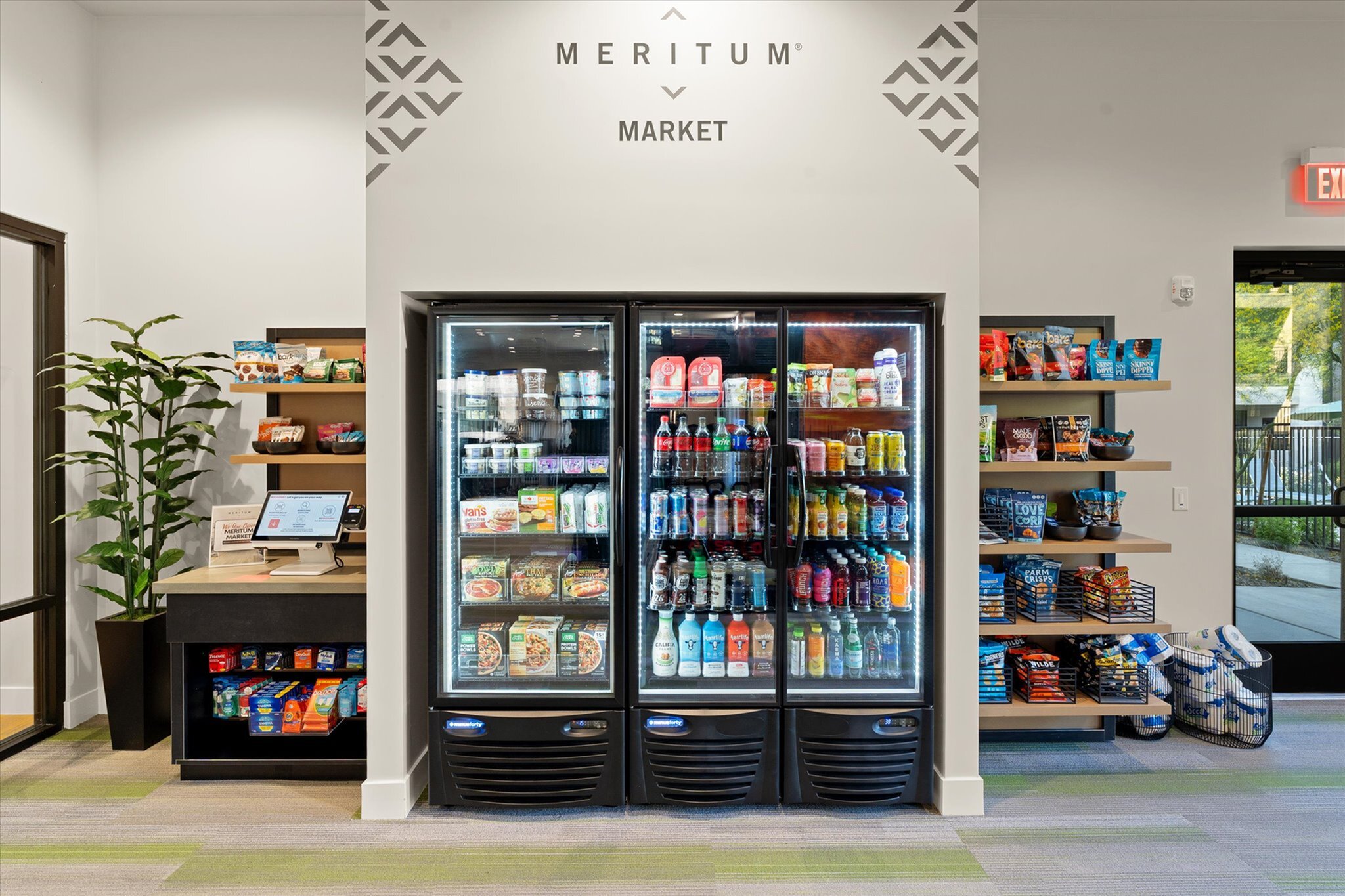 Modern community market with well-stocked refrigerated drinks and snacks section, stylish interior design, and convenient self-checkout kiosk.
