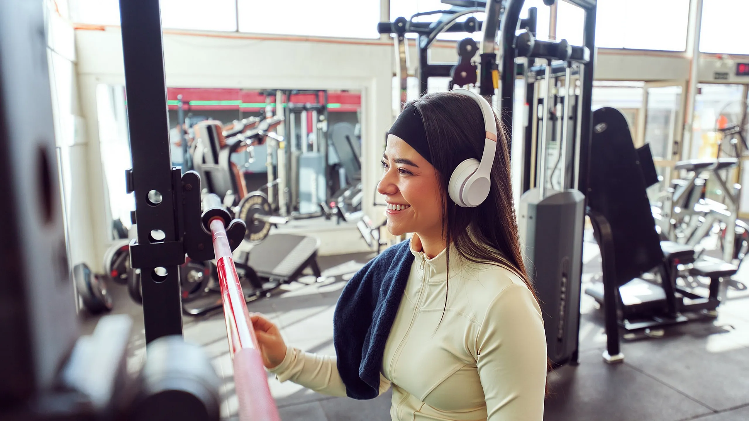 Energetic woman in modern gym, smiling while using a squat rack, wearing headphones and sporty attire.