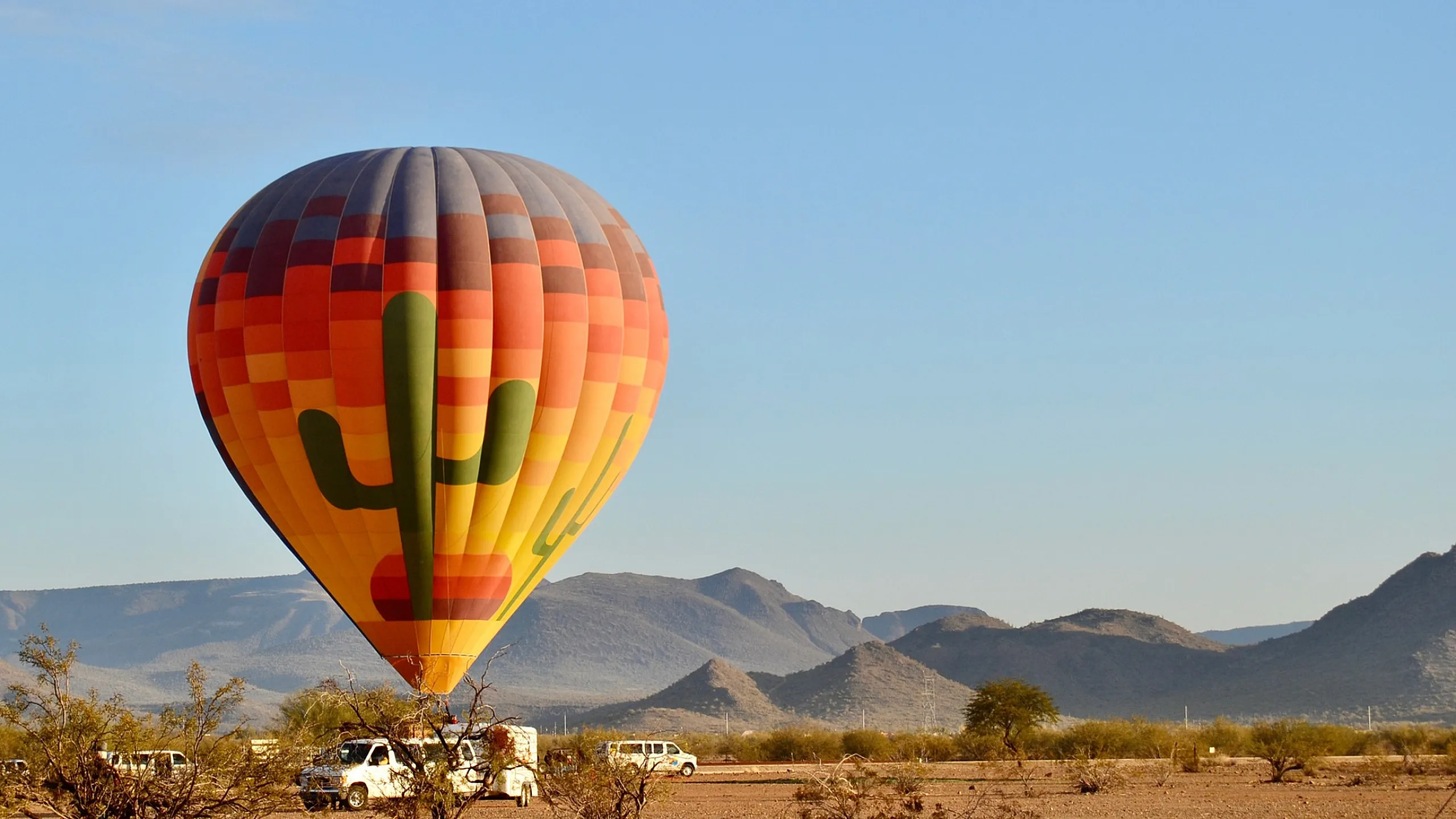 Vibrant hot air balloon with cactus design floating above a scenic desert landscape and distant mountains under a clear blue sky.