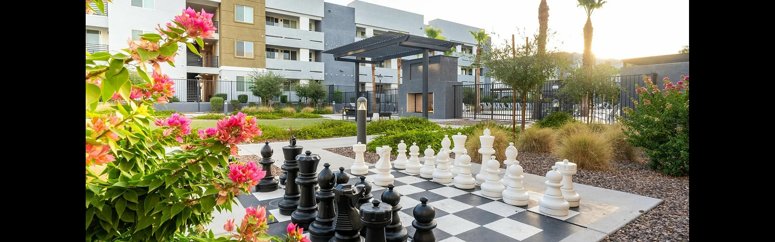 Vibrant apartment view with lush landscaping, a life-sized outdoor chess set, and modern architecture under a sunny sky.
