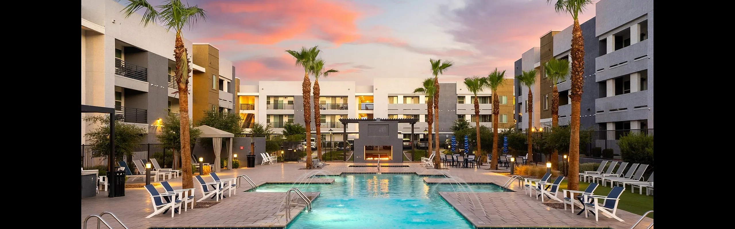 Luxurious apartment complex with a stunning sunset backdrop, featuring a sparkling outdoor pool surrounded by palm trees and modern architecture.