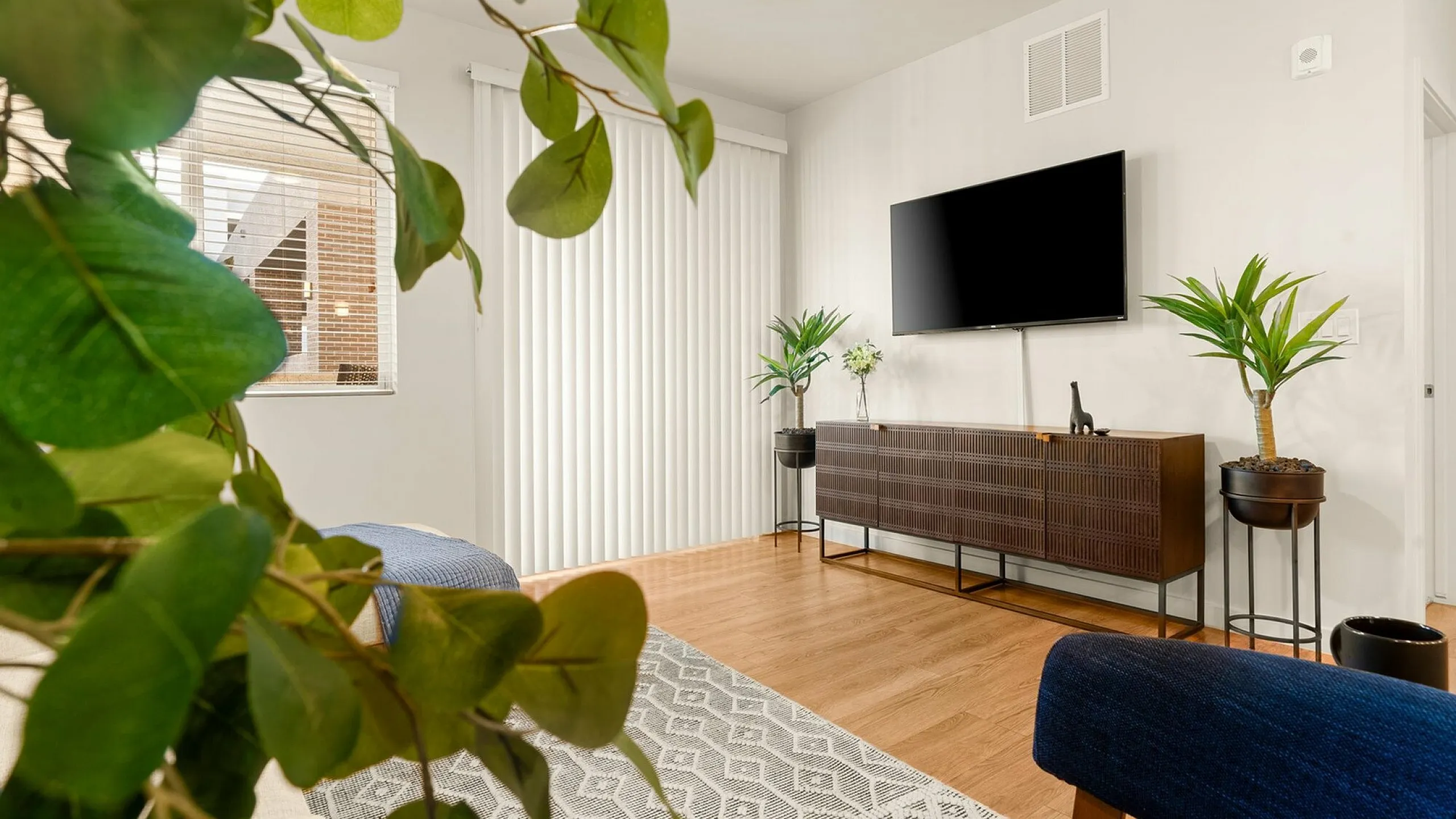 Inviting living room with modern decor featuring a sleek TV mounted on the wall, stylish wooden console, lush plants, and cozy seating, perfect for relaxation.