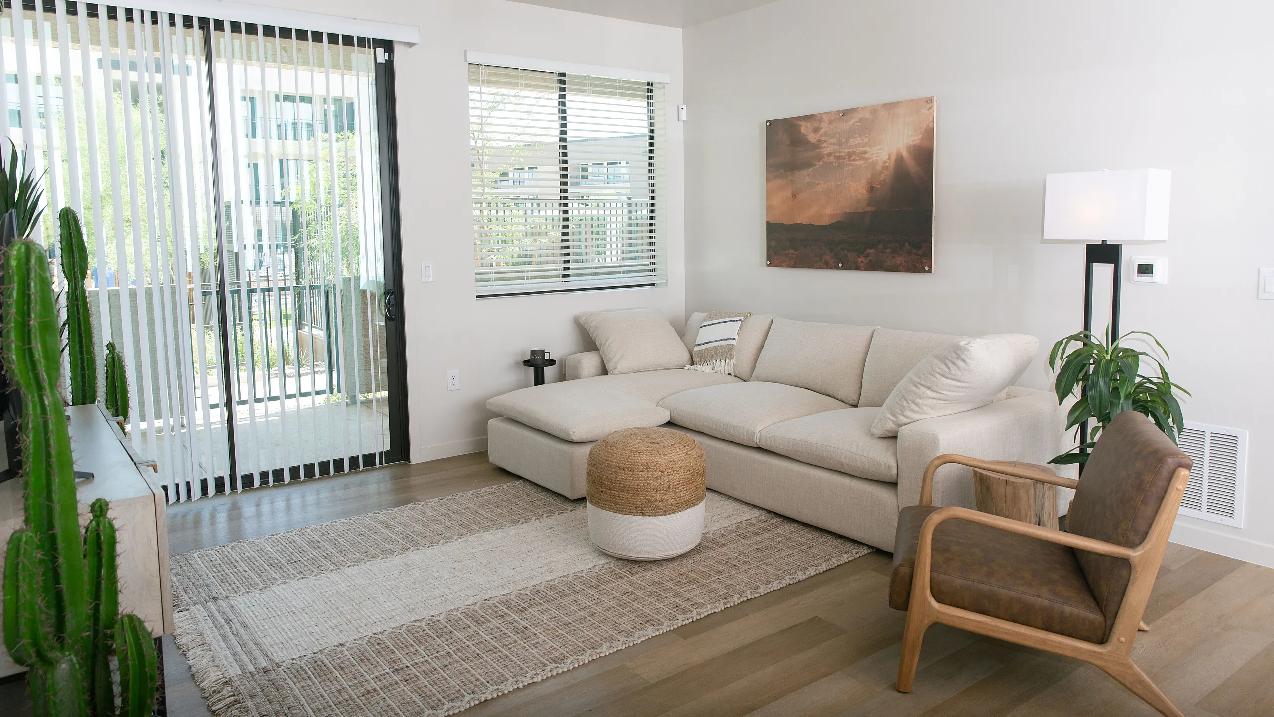 Modern living room with cozy beige sectional sofa, stylish armchair, and natural light streaming through large sliding doors. Elegant decor includes a potted cactus, artistic wall art, and a sleek floor lamp, creating a warm and inviting at