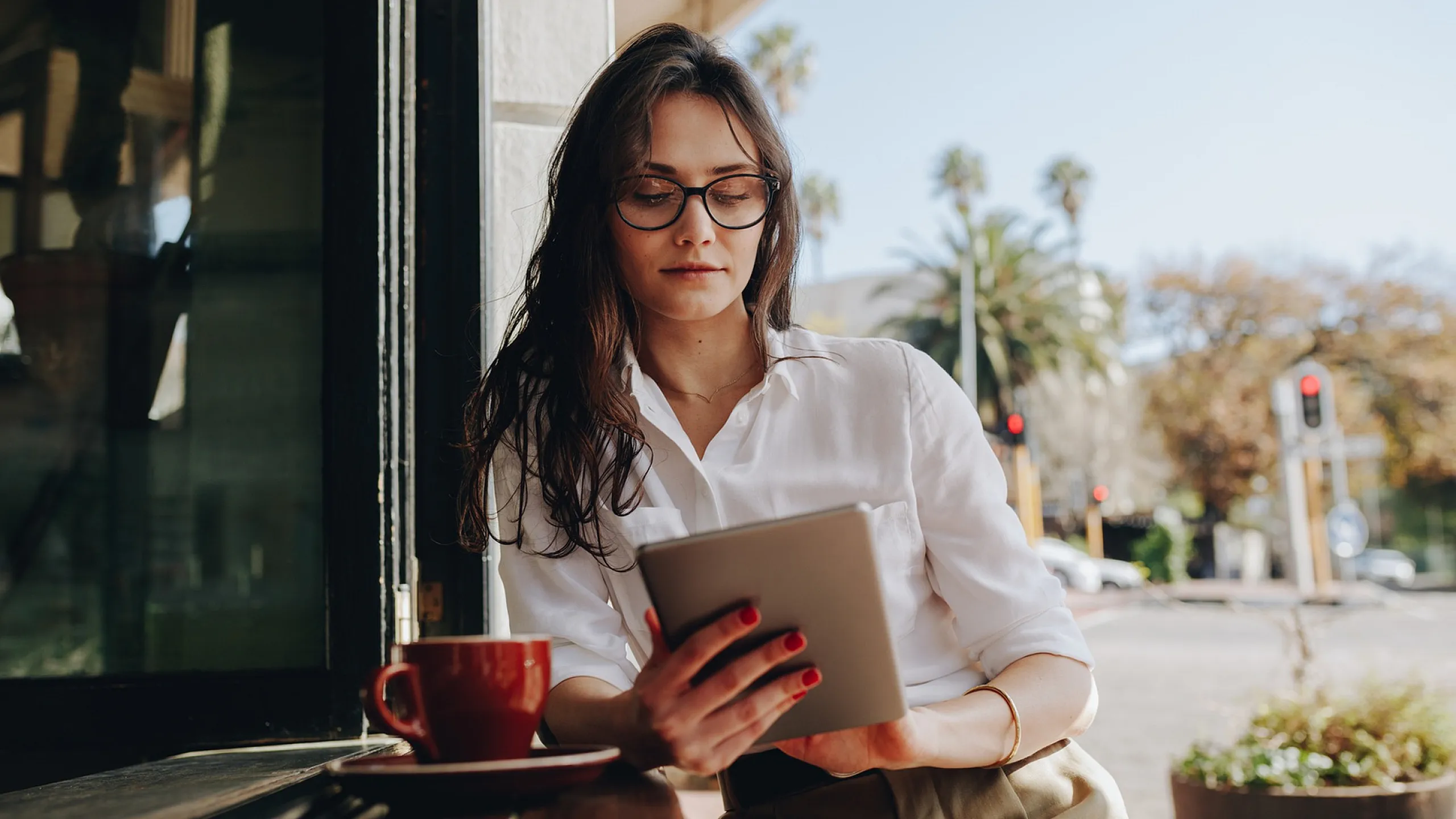 Woman reading on a tablet in a cozy caf&eacute;, surrounded by lush palm trees and sunlight.