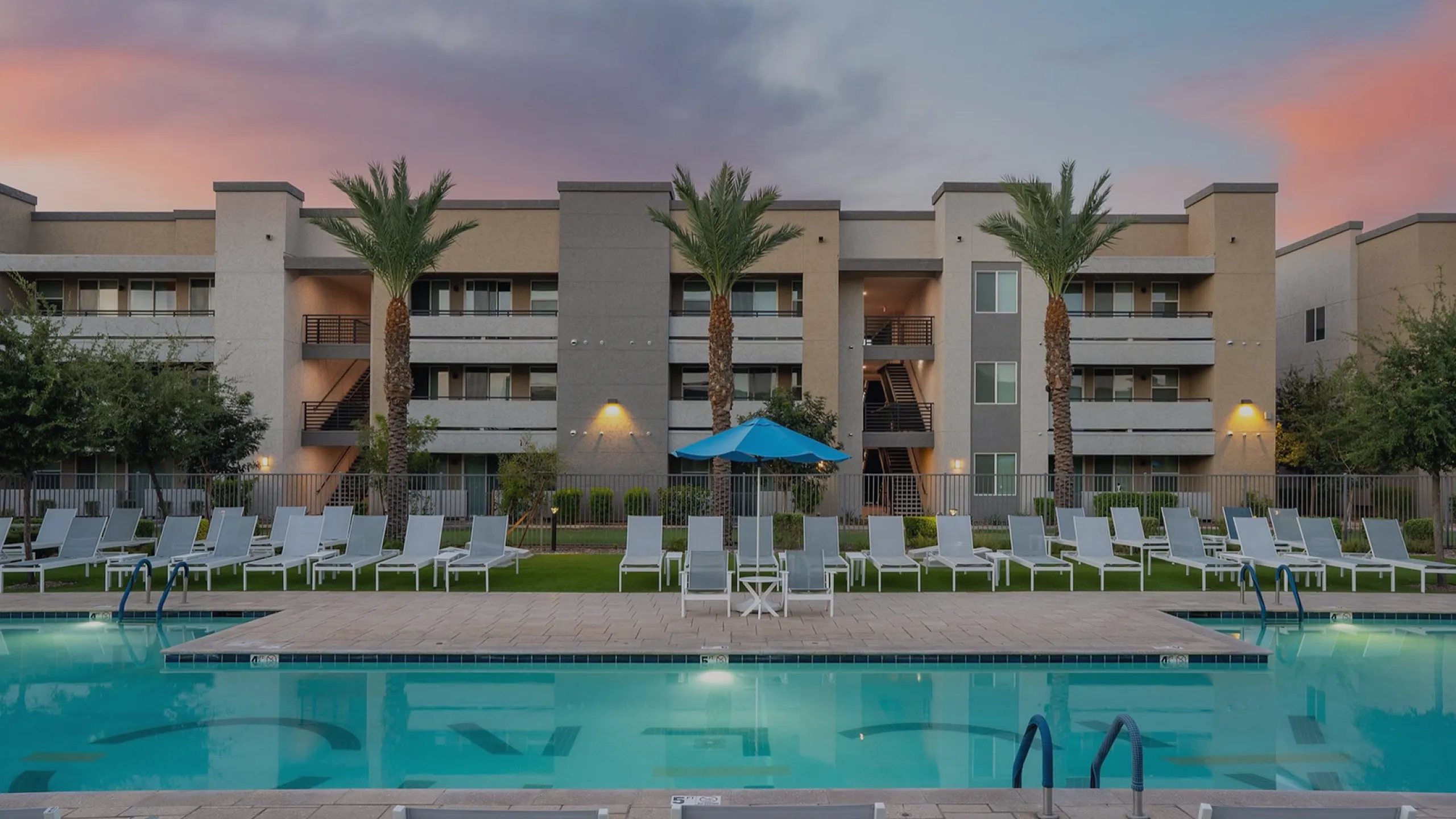 Modern apartment complex with a luxurious poolside view, showcasing a serene sunset backdrop and palm trees.