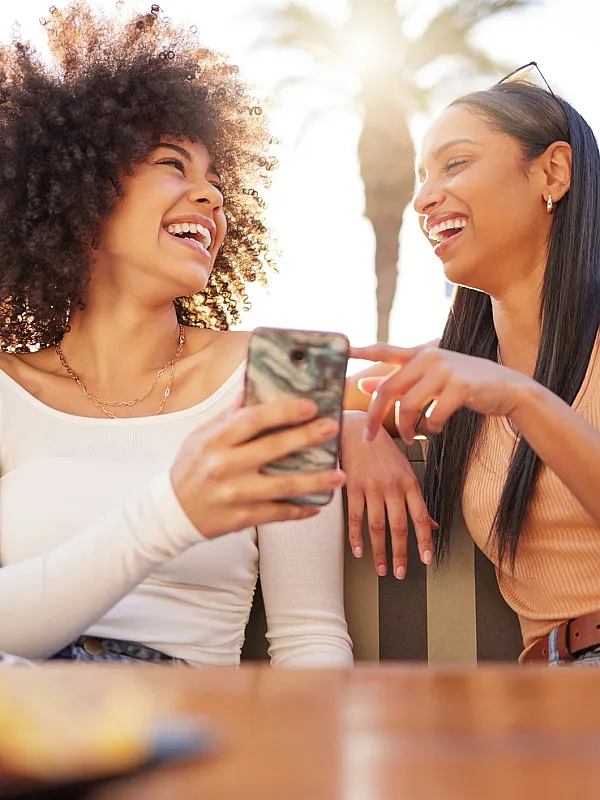 Two friends enjoying a sunny day outdoors, laughing and sharing a moment while looking at a smartphone.