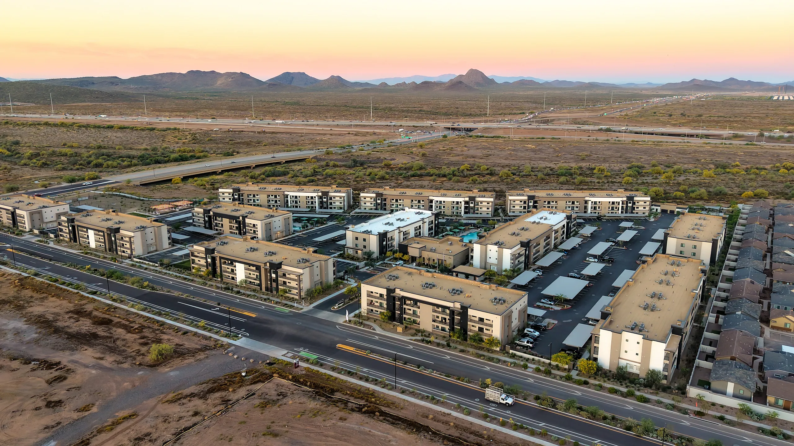 Aerial view of a modern apartment complex surrounded by scenic desert landscape and distant mountains at sunset, highlighting accessibility and serene living environment.