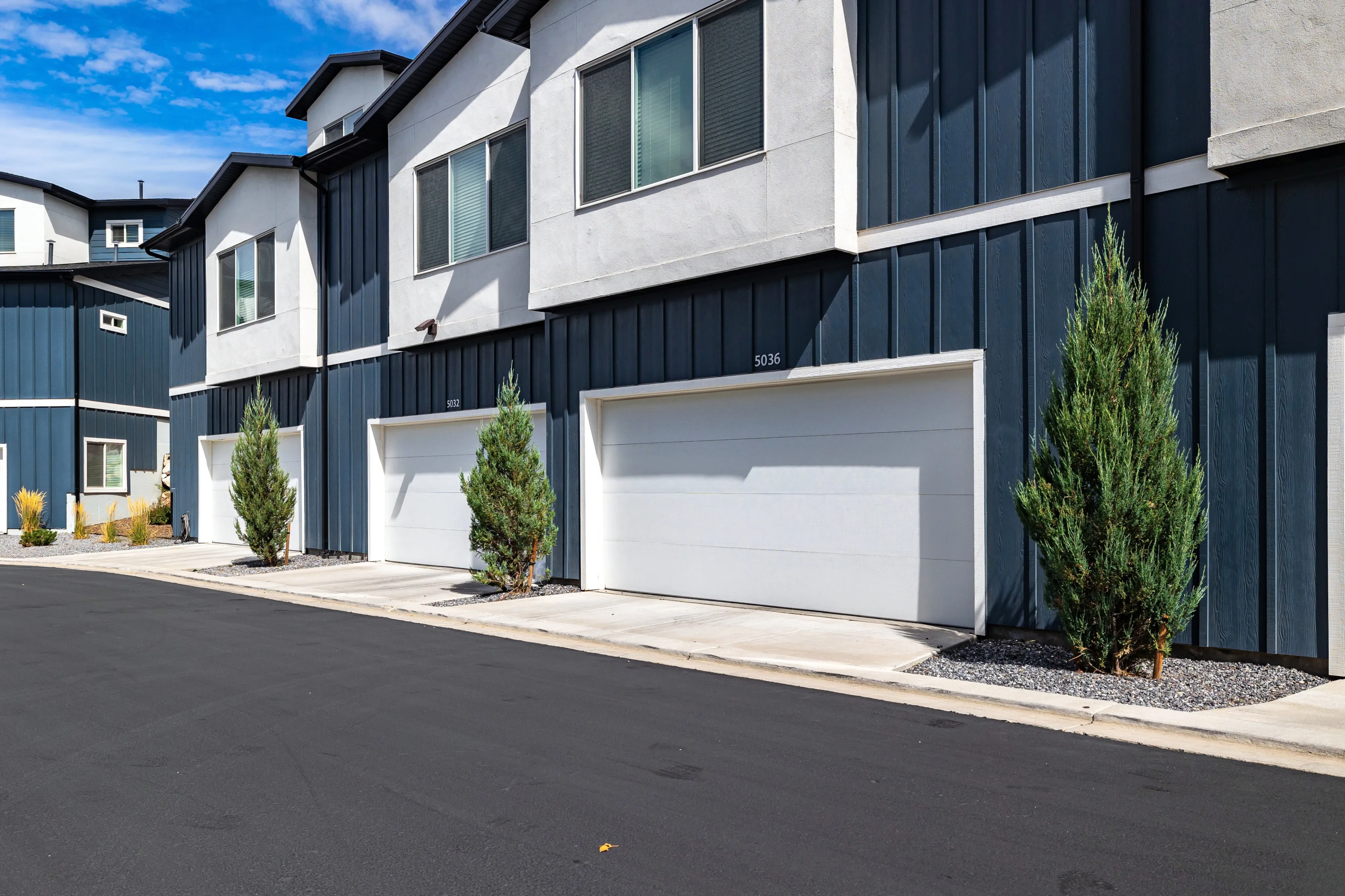 Modern townhouse exterior featuring sleek design with white and dark blue siding, large windows, and dual garage doors. Beautifully landscaped with evergreen shrubs lining the sidewalk.