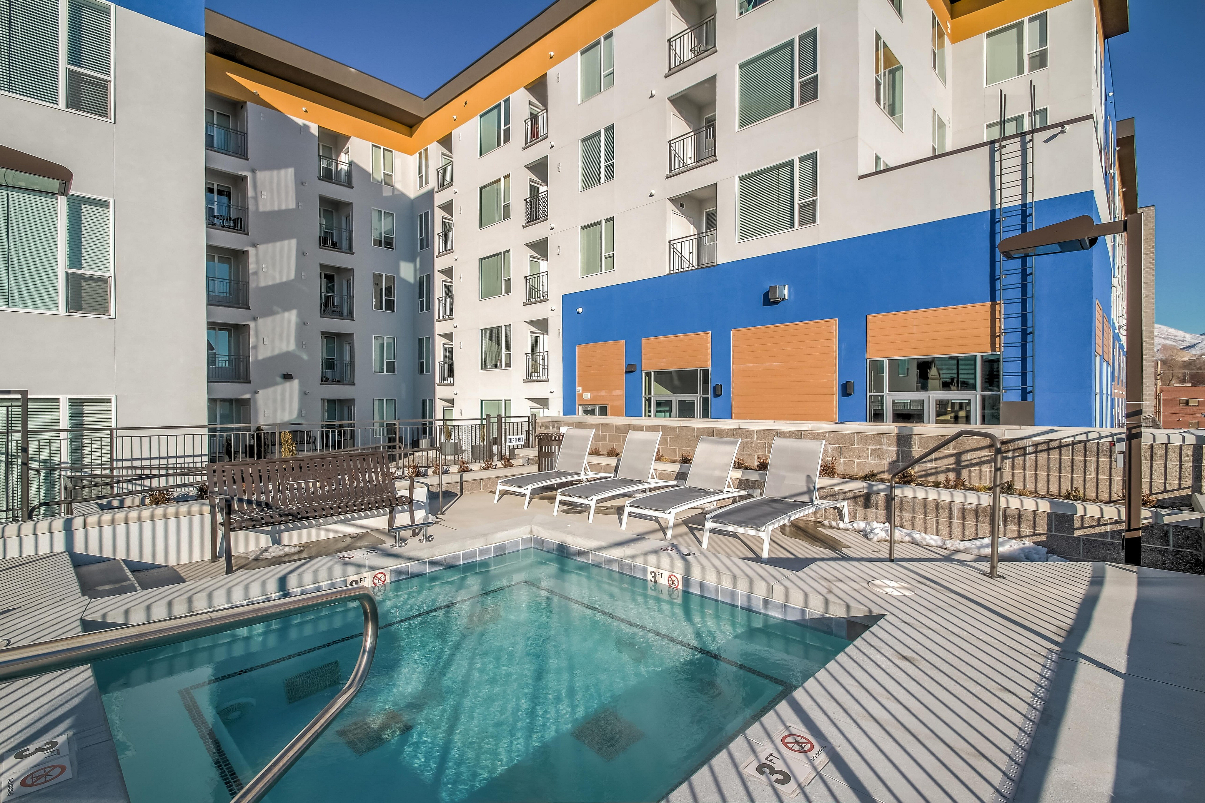 Modern apartment complex featuring a sunlit pool area with stylish loungers, set against a vibrant blue and white facade.