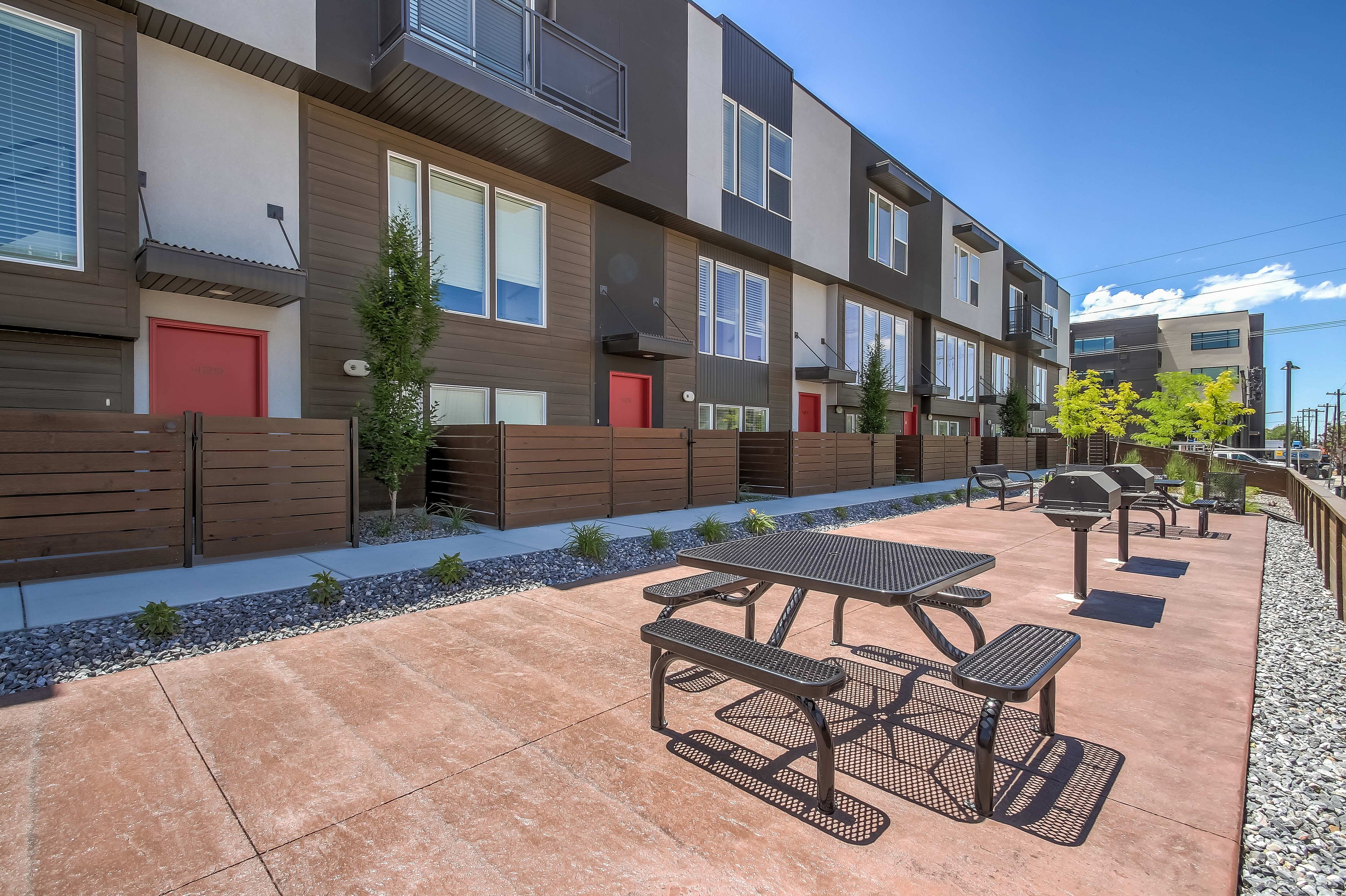 Modern townhouse exterior with stylish red doors, private fenced patios, and outdoor seating on a sunlit terrace.