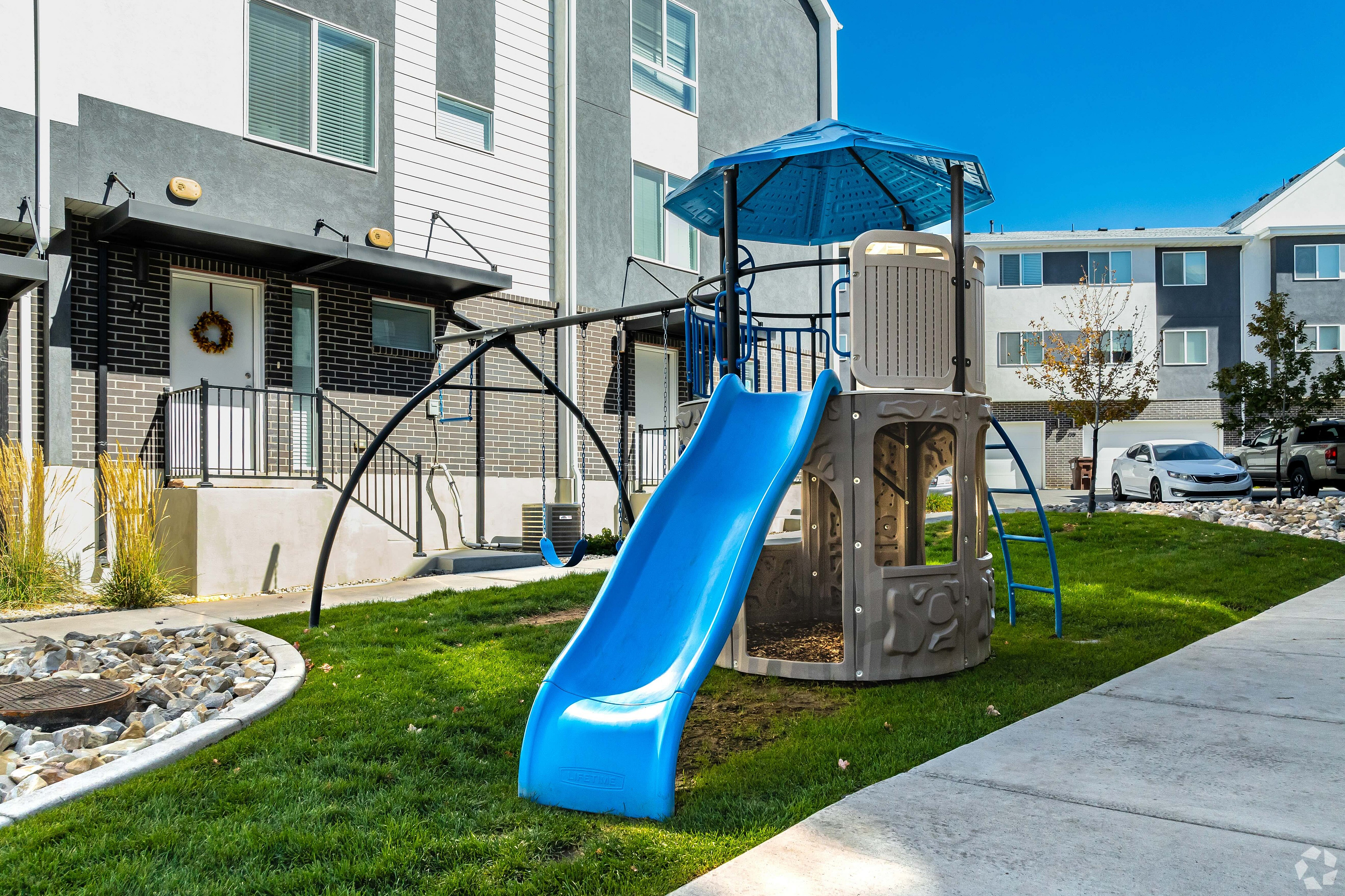 Charming residential area featuring a colorful playground with slide, surrounded by modern townhouses and lush green lawn, ideal for family living.