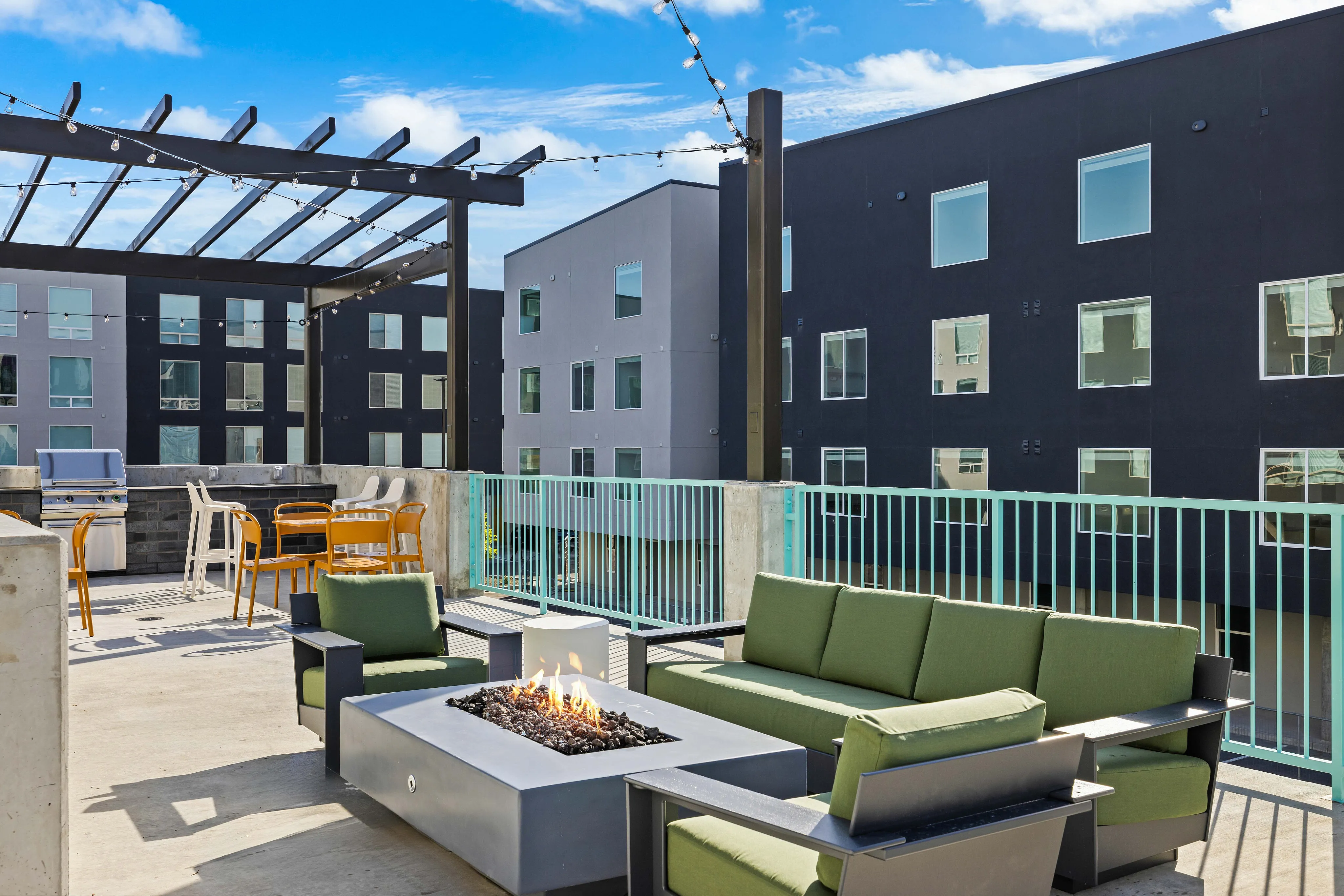Modern outdoor patio with cozy green seating, fire pit, and BBQ grill, perfect for entertaining against a backdrop of stylish apartment buildings.