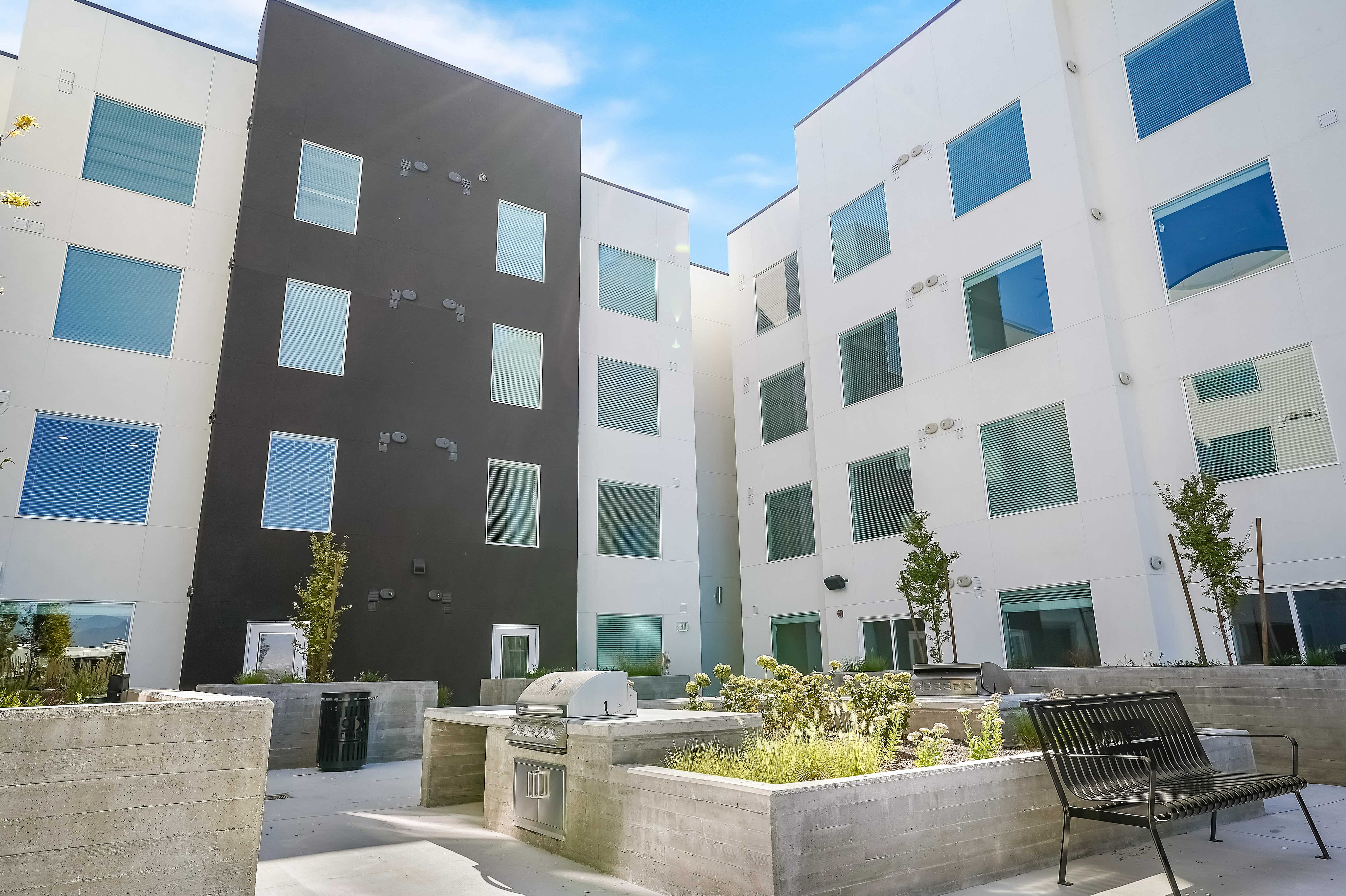 Modern apartment complex courtyard featuring sleek architecture, abundant windows, and a communal BBQ area with landscaped greenery.