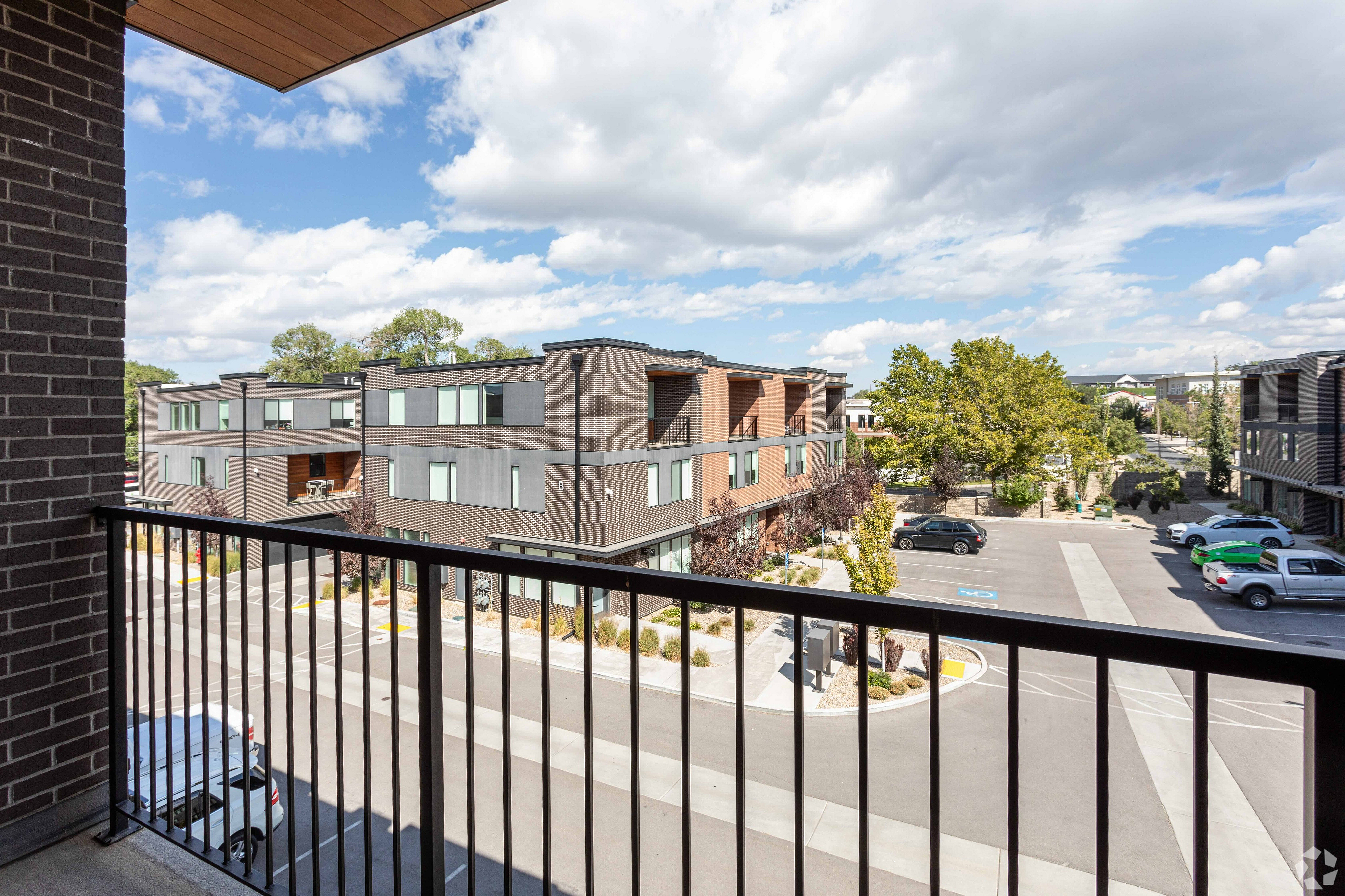 Modern apartment balcony view showcasing stylish architecture, lush greenery, and a serene parking area under a bright blue sky.