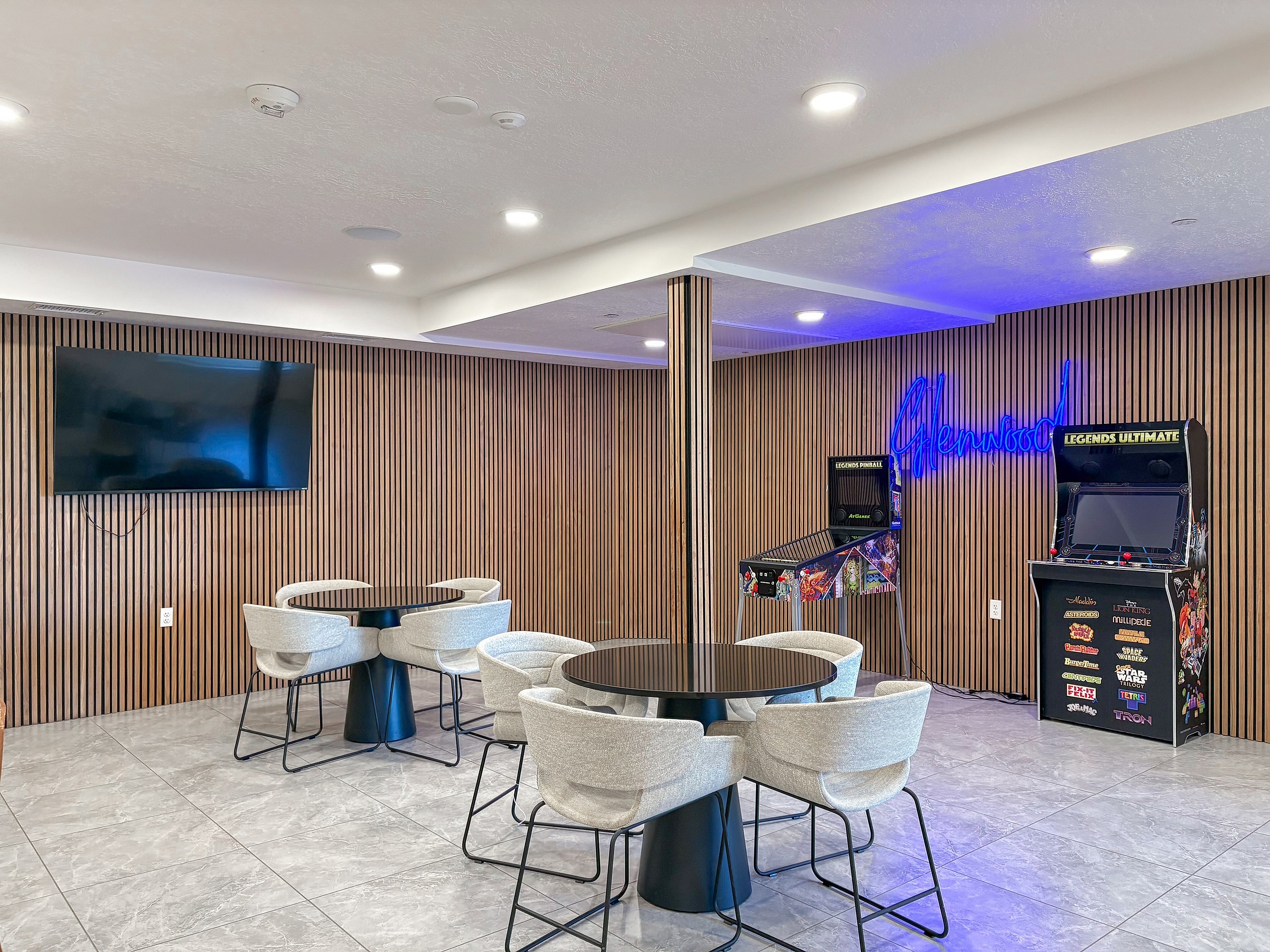 Modern entertainment room featuring sleek arcade machines, stylish lounge chairs, a flat-screen TV, and elegant wood paneling.