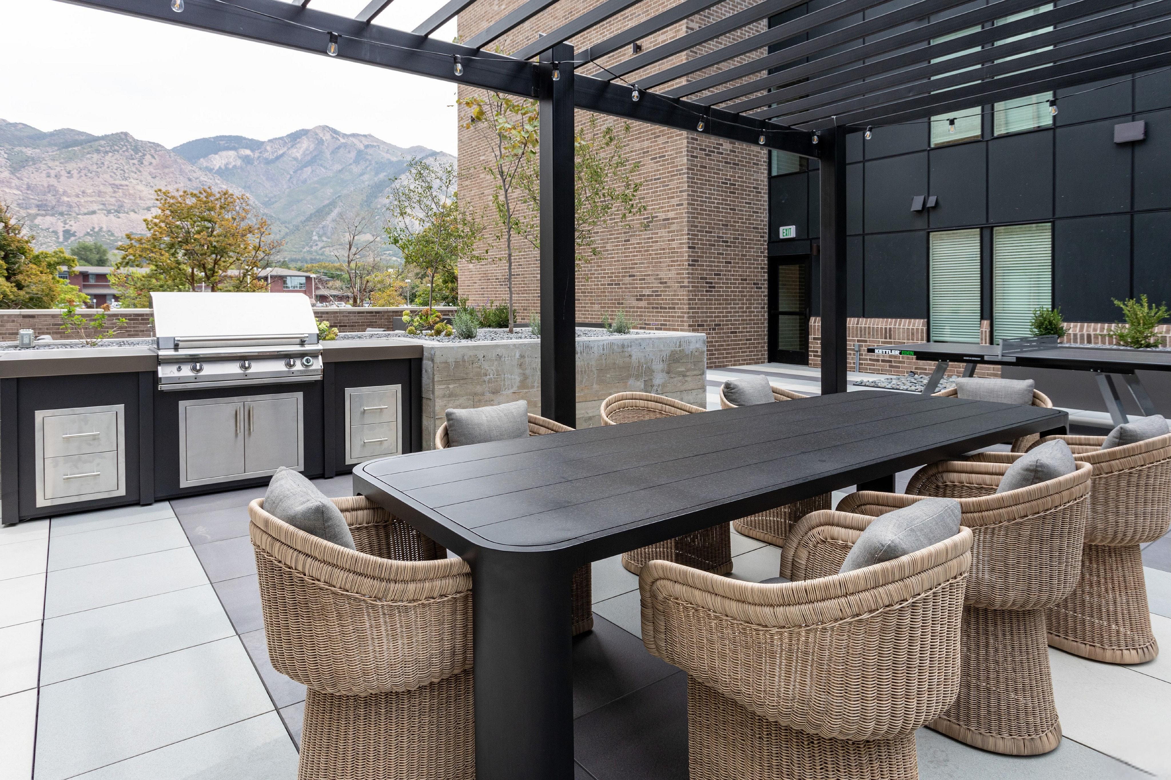 Elegant outdoor terrace with a sleek grill, modern black dining table, and inviting wicker chairs, set against a backdrop of stunning mountain views.