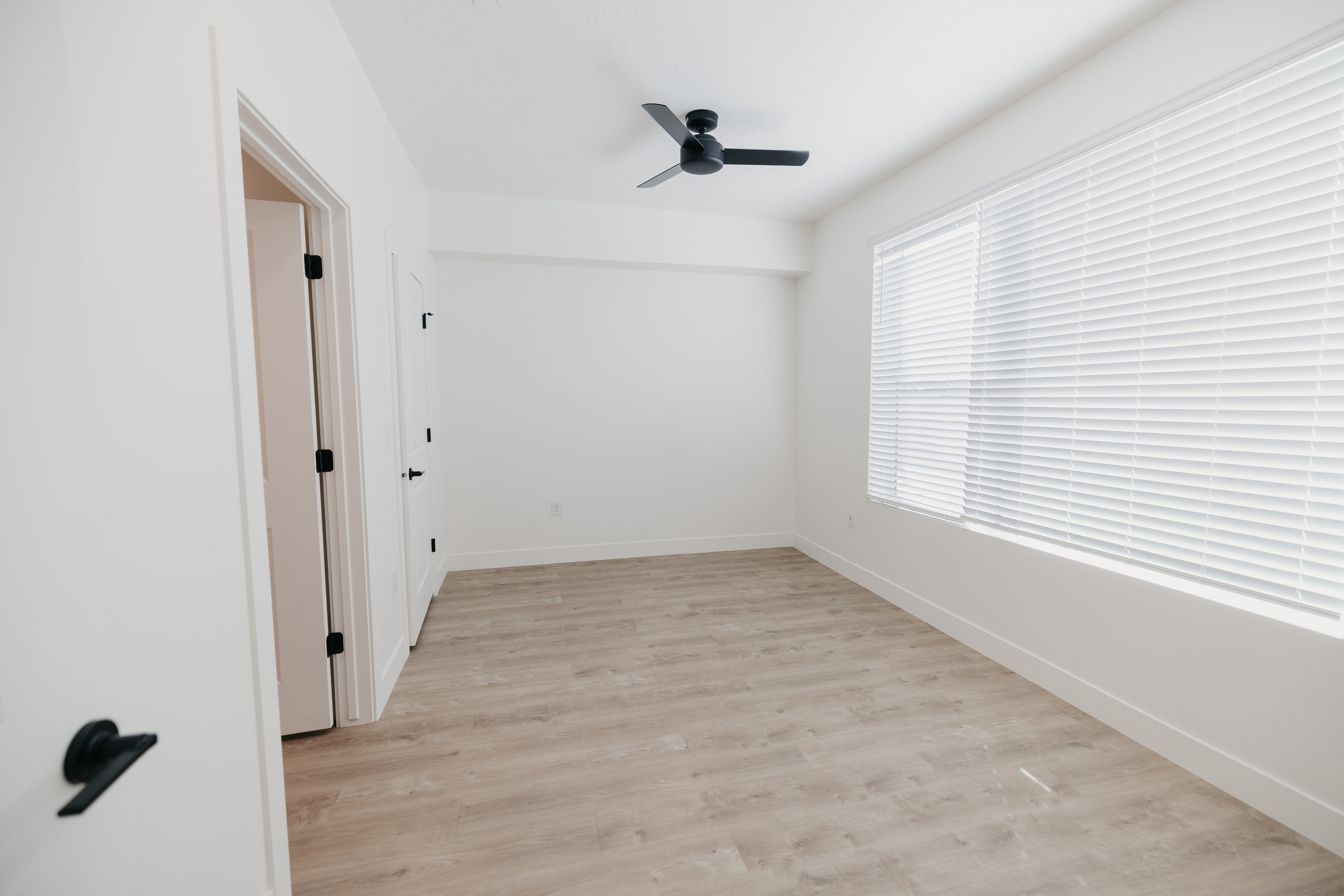 Bright and airy unfurnished room with sleek wooden flooring, modern ceiling fan, and large window with blinds for natural light.
