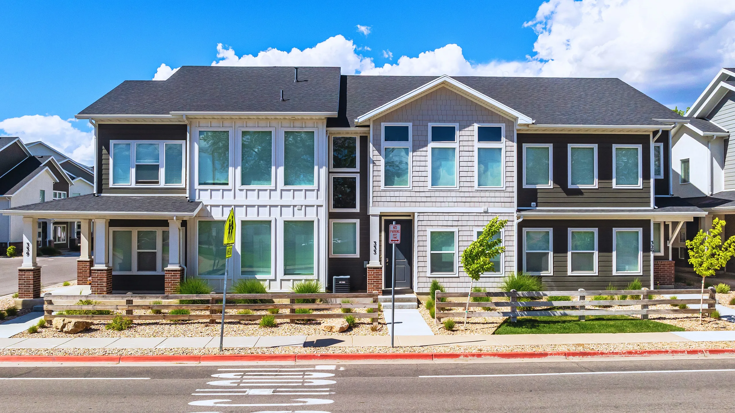 Modern two-story duplex with white and gray siding, featuring large windows and a spacious front yard under a bright blue sky.