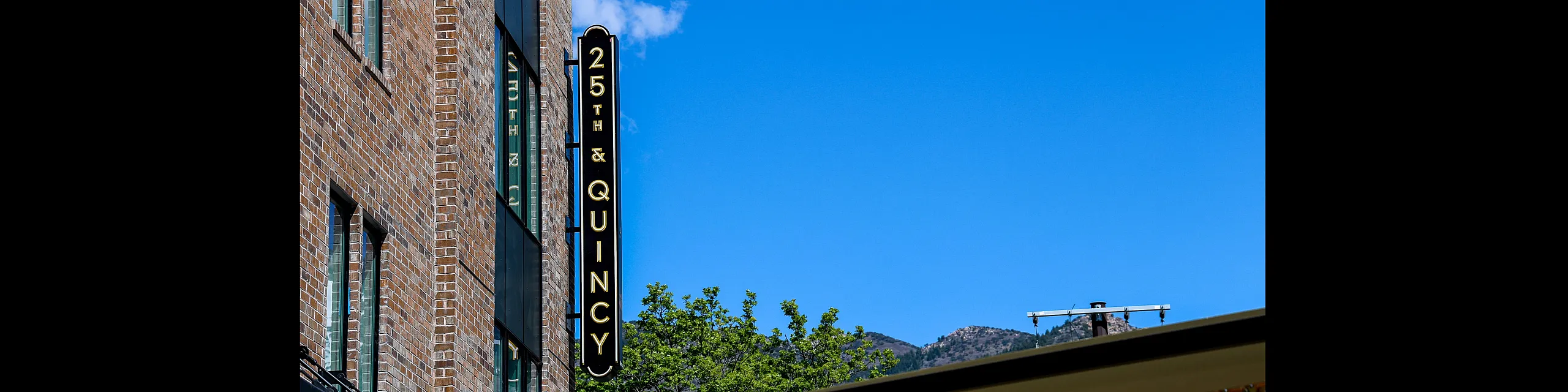 Charming urban apartment facade with a bold 25th & Quincy sign against a bright blue sky, offering a blend of modern city living and scenic mountain views.