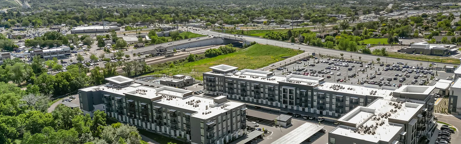 Aerial view of a modern apartment complex surrounded by lush greenery and majestic mountains, showcasing ample parking and convenient location near urban amenities.