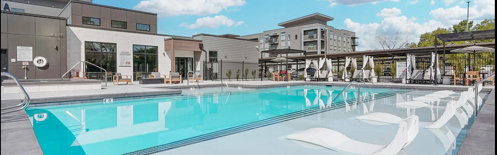 Luxurious apartment complex featuring a sparkling swimming pool with lounge chairs, cabanas, and a modern building backdrop under a vibrant blue sky.