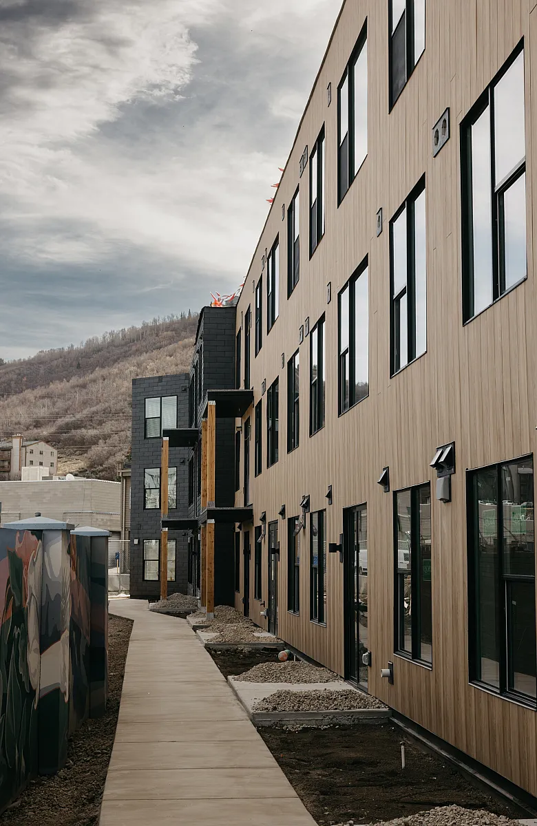 Modern apartment exterior with wood paneling and large windows, set against a picturesque mountain backdrop and vibrant mural art along the walkway.
