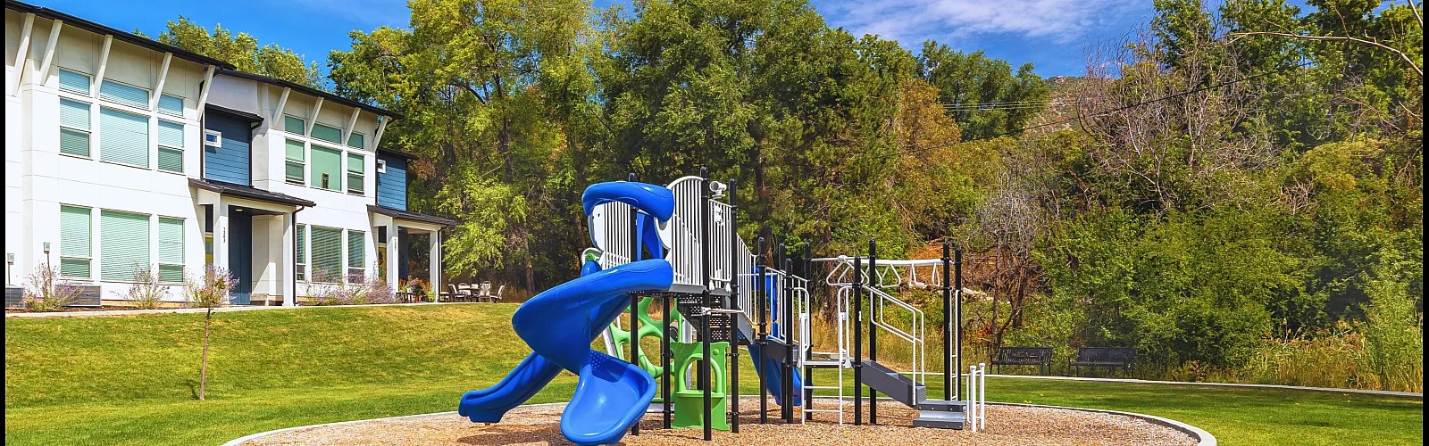 Charming community playground with vibrant blue slides surrounded by lush greenery and a modern building backdrop.