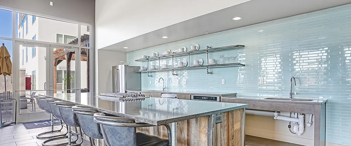 Modern kitchen featuring a sleek island with metallic stools, soft blue tile backsplash, and ample natural light from large windows.