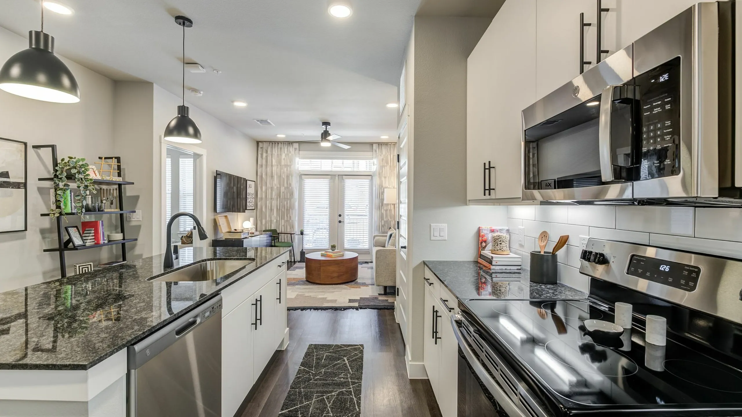 Modern kitchen with sleek black countertops, stainless steel appliances, and stylish pendant lighting, leading into a cozy, open-concept living room with natural light.