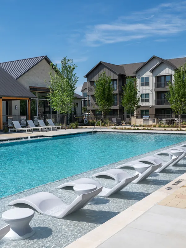 Luxurious apartment complex with a sparkling swimming pool, modern poolside loungers, and lush landscaping under a clear blue sky.