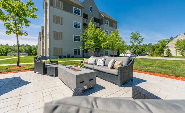 Inviting outdoor lounge area with modern seating and fire pit in front of a contemporary apartment building surrounded by lush green lawns and trees.