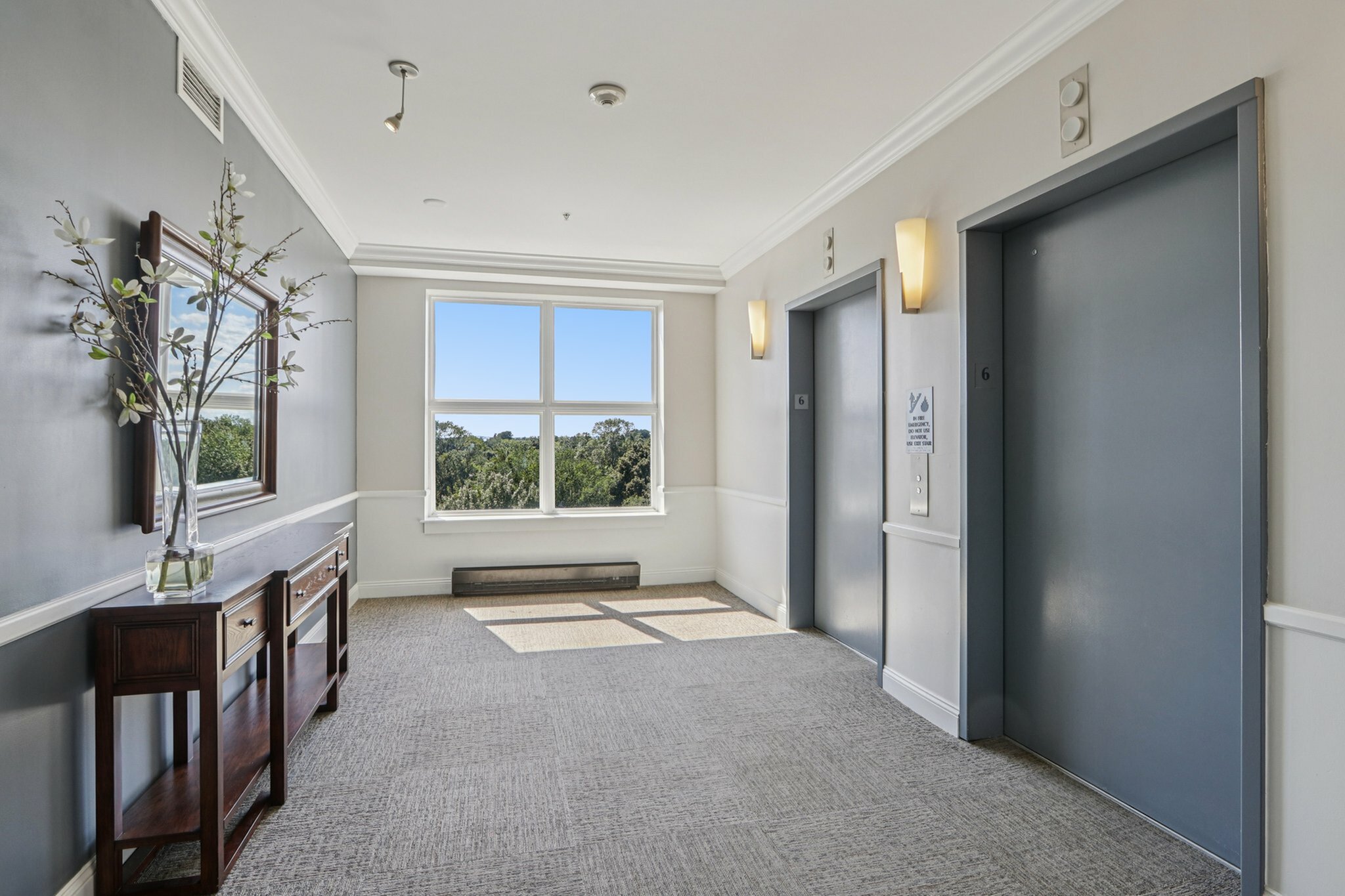 Bright and modern apartment corridor featuring elegant elevators, a large window with scenic views, and sophisticated decor.