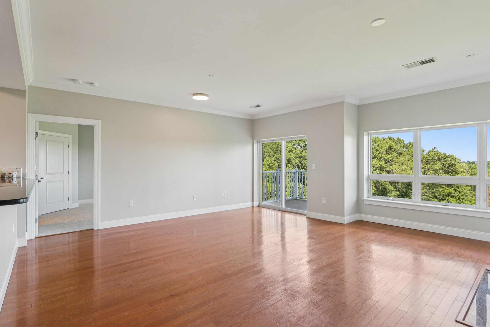 Bright and spacious living room with polished hardwood floors, large windows overlooking lush greenery, and a balcony for outdoor enjoyment.