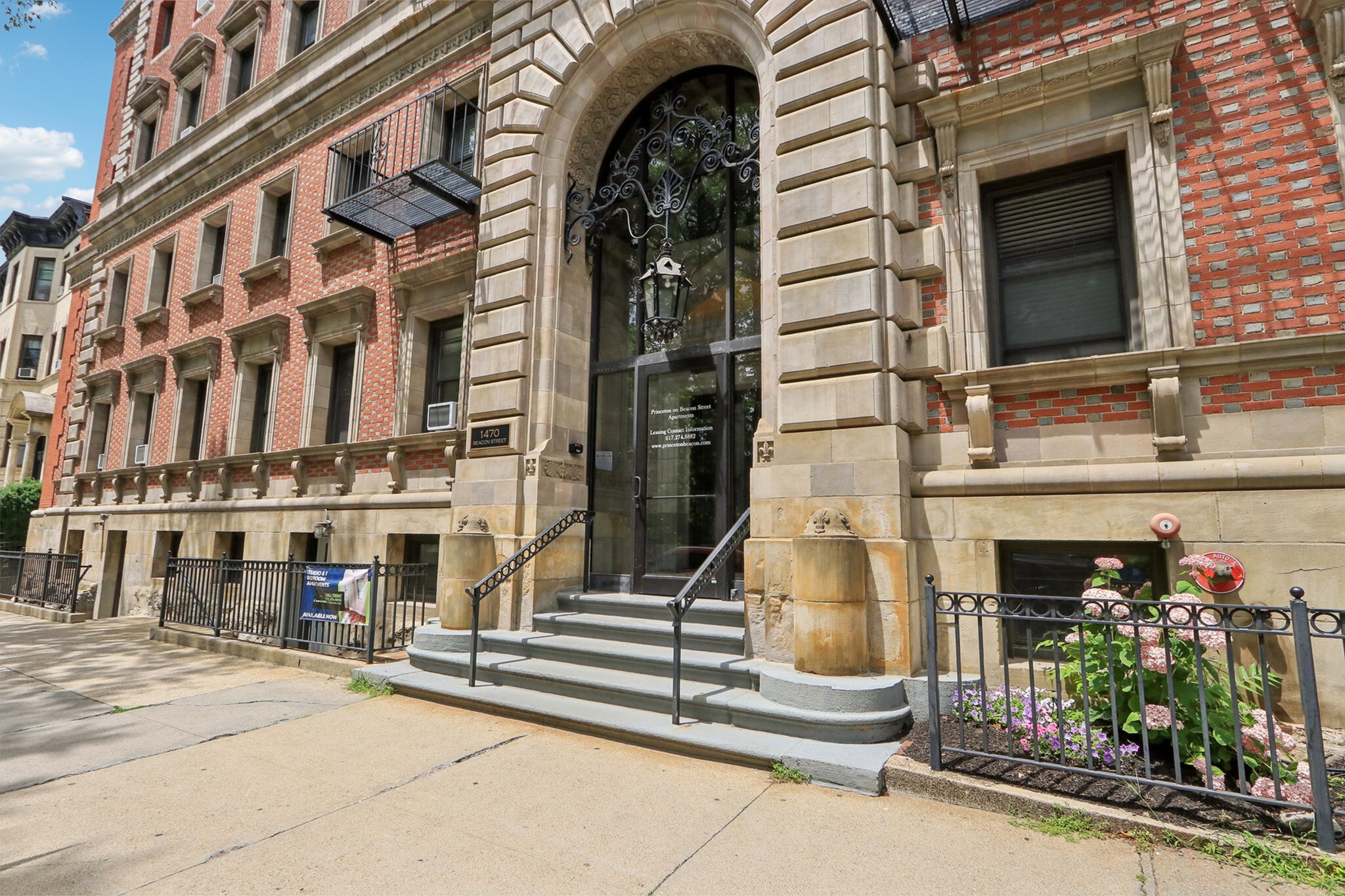 Elegant historic building entrance with ornate archway and classic brickwork, featuring charming ironwork and vibrant flower bed, showcasing timeless architectural beauty.