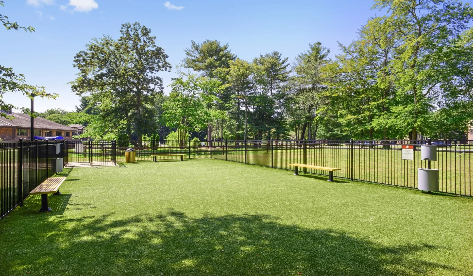Inviting outdoor space featuring a fenced-in grassy area with benches, surrounded by lush trees under a clear blue sky.
